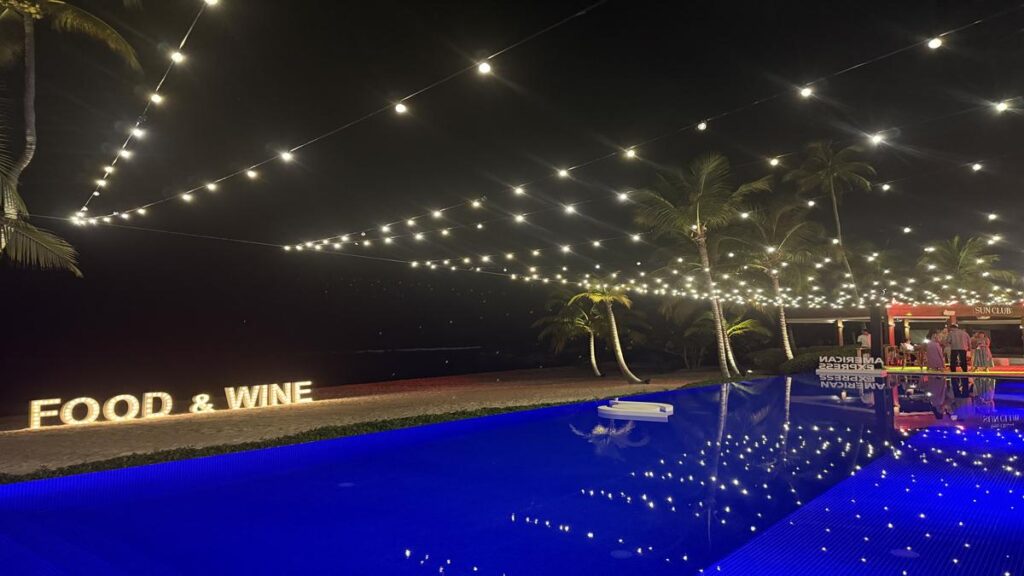 Blue illuminated swimming pool with Food & Wine sign and string lights at Minitas Beach Club