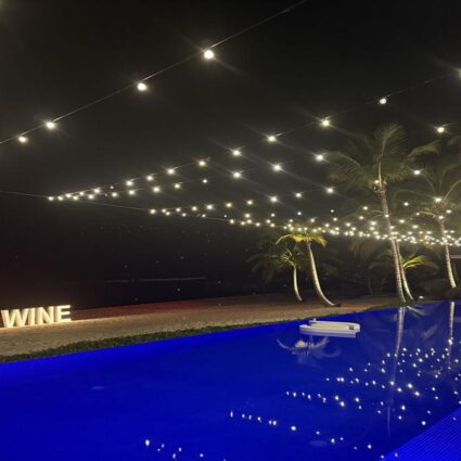 Blue illuminated swimming pool with Food & Wine sign and string lights at Minitas Beach Club